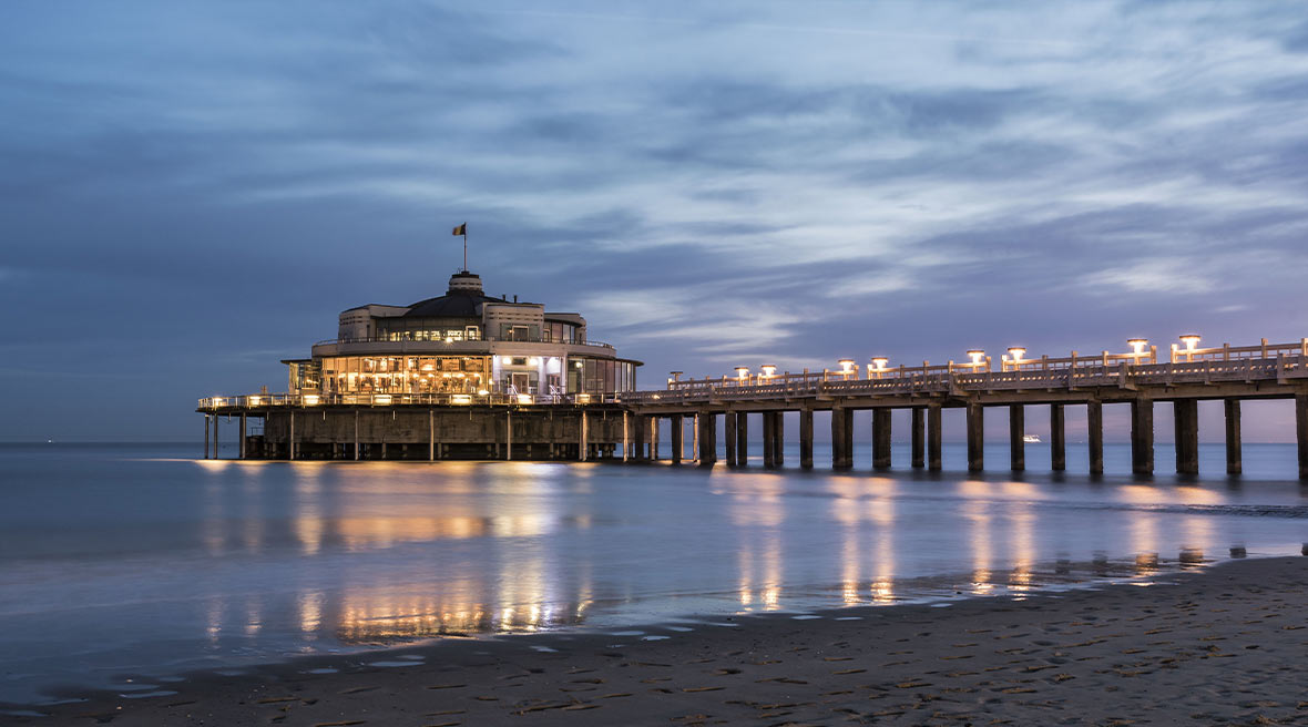 A lit up pier and building at sunrise with sand and sea in the foreground